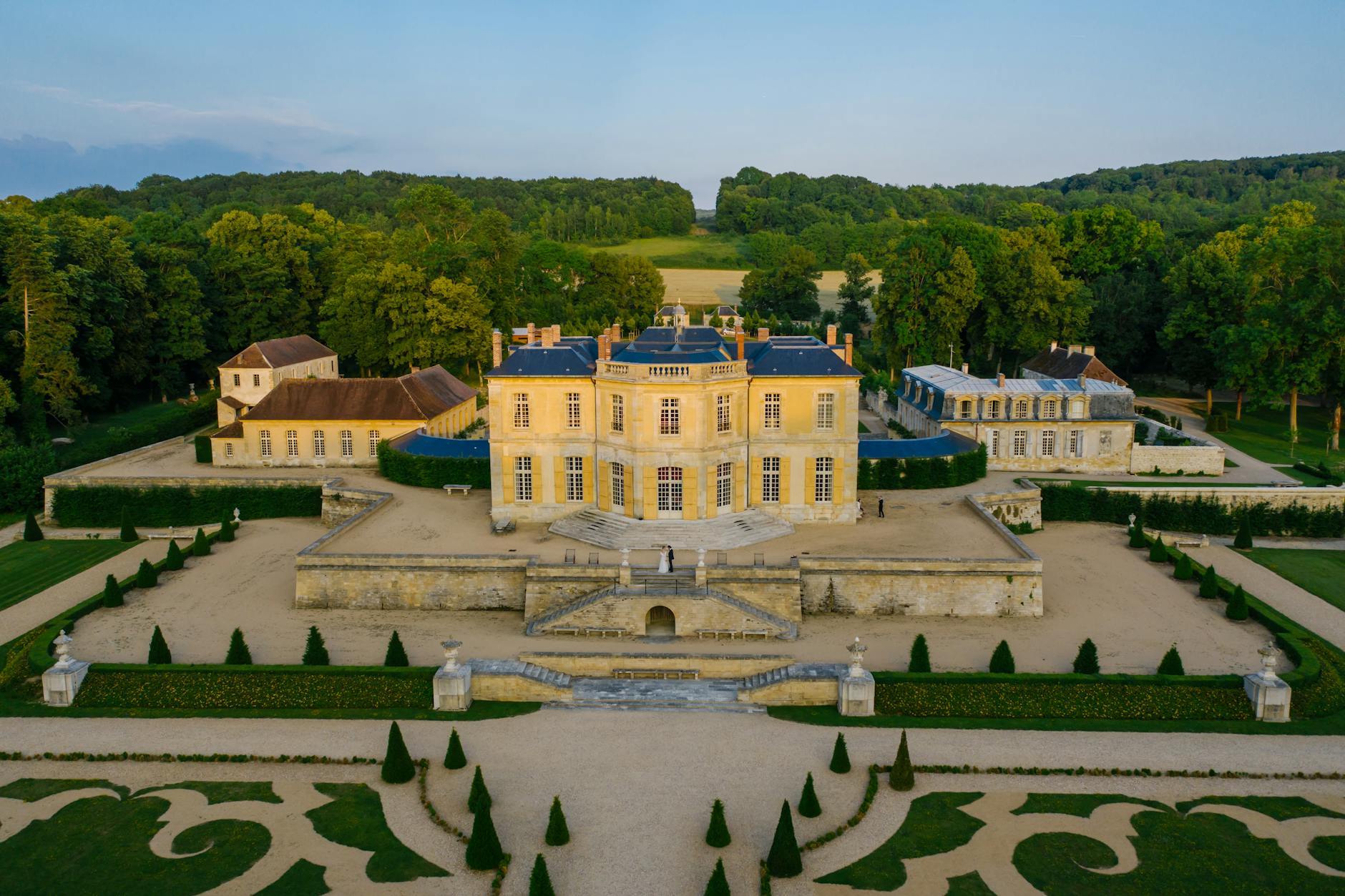 Elegant aerial view of a historic French château surrounded by lush gardens and trees, in summer.