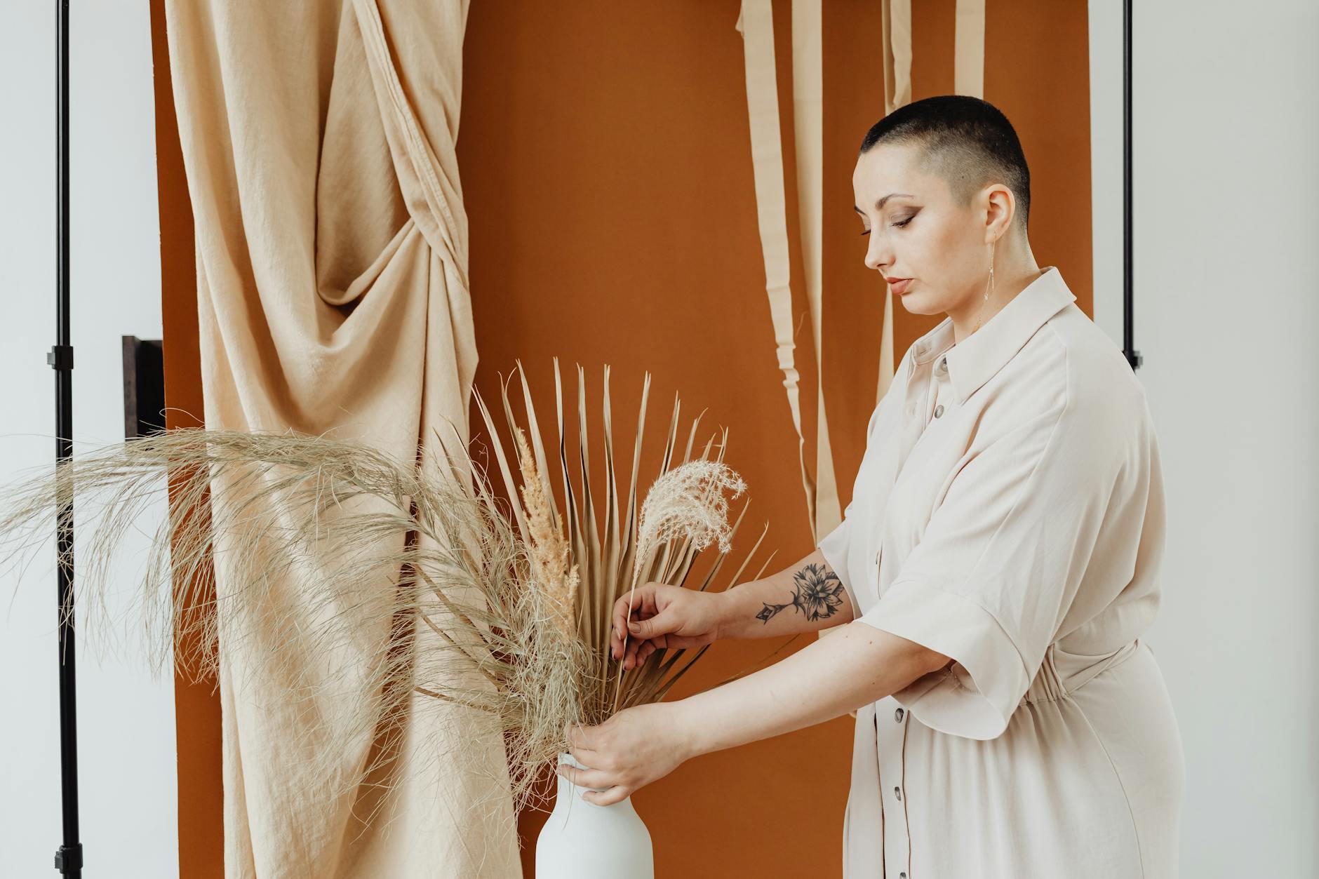 A stylish woman arranging dried flowers in a studio with a warm beige backdrop.