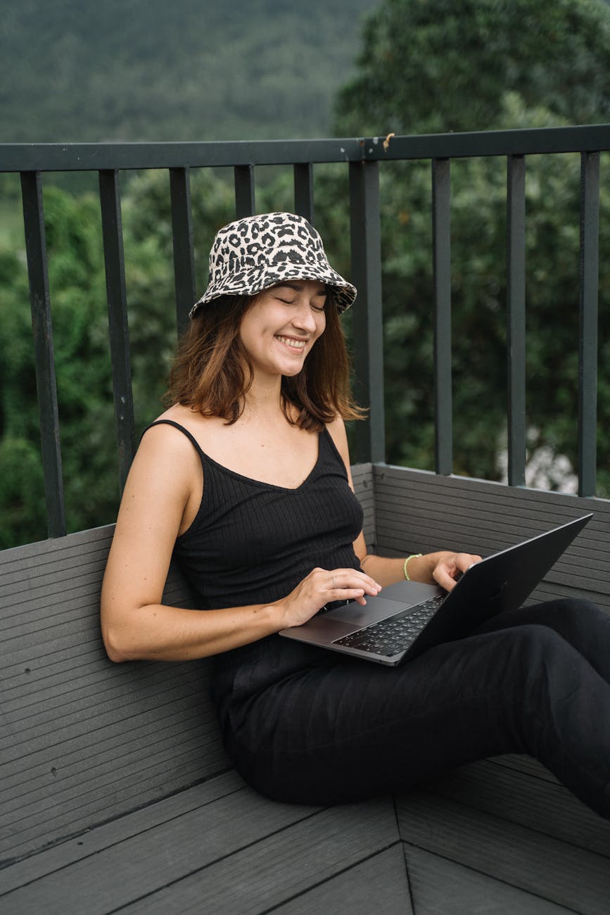Smiling woman in a leopard hat and black outfit working on a laptop outdoors.